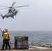 A military helicopter is hovering above the flight deck of a naval vessel at sea, while two deck crew members in safety gear appear to be preparing for a fueling or landing operation. One crew member in a yellow vest is holding what appears to be a grounding wand or fuel hose toward the helicopter. A metal equipment unit with orange hoses is positioned on the deck nearby.
