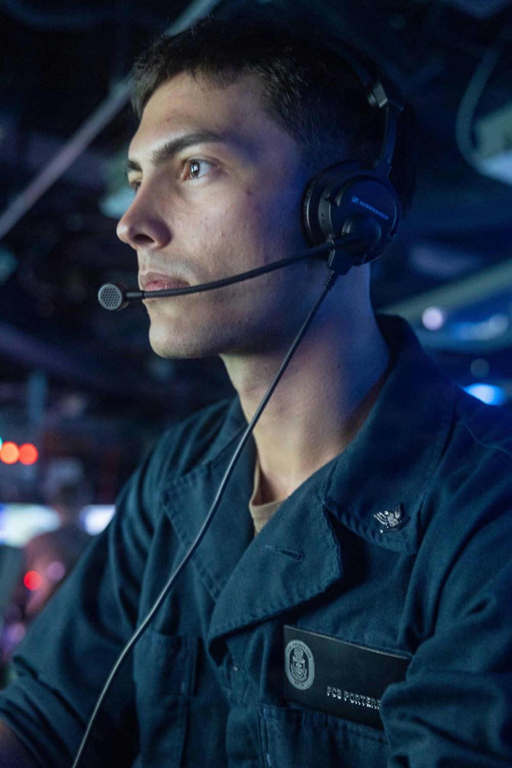 A U.S. Navy sailor wearing a Sennheiser communications headset is photographed in a dimly lit combat information center or operations room aboard a naval vessel. The sailor is wearing a dark blue Navy working uniform with a name tag reading 'FC3 PORTER' and a ship's crest badge visible on the chest. The blue-tinted lighting and background displays suggest an active operational environment.