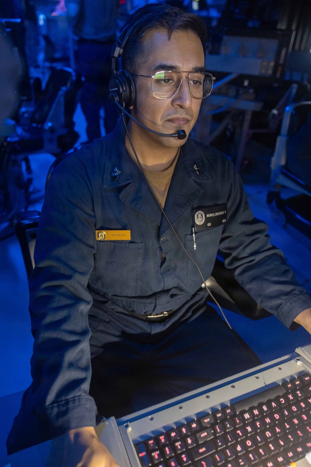 A U.S. Navy sailor in dark blue coveralls (NWU Type III or similar working uniform) is seated at a workstation in what appears to be a combat information center or operations room aboard a naval vessel. The individual is wearing a communications headset with boom microphone and glasses, actively monitoring equipment. The environment is illuminated with blue ambient lighting, typical of tactical operations spaces.