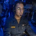 A U.S. Navy sailor in dark blue coveralls (NWU Type III or similar working uniform) is seated at a workstation in what appears to be a combat information center or operations room aboard a naval vessel. The individual is wearing a communications headset with boom microphone and glasses, actively monitoring equipment. The environment is illuminated with blue ambient lighting, typical of tactical operations spaces.