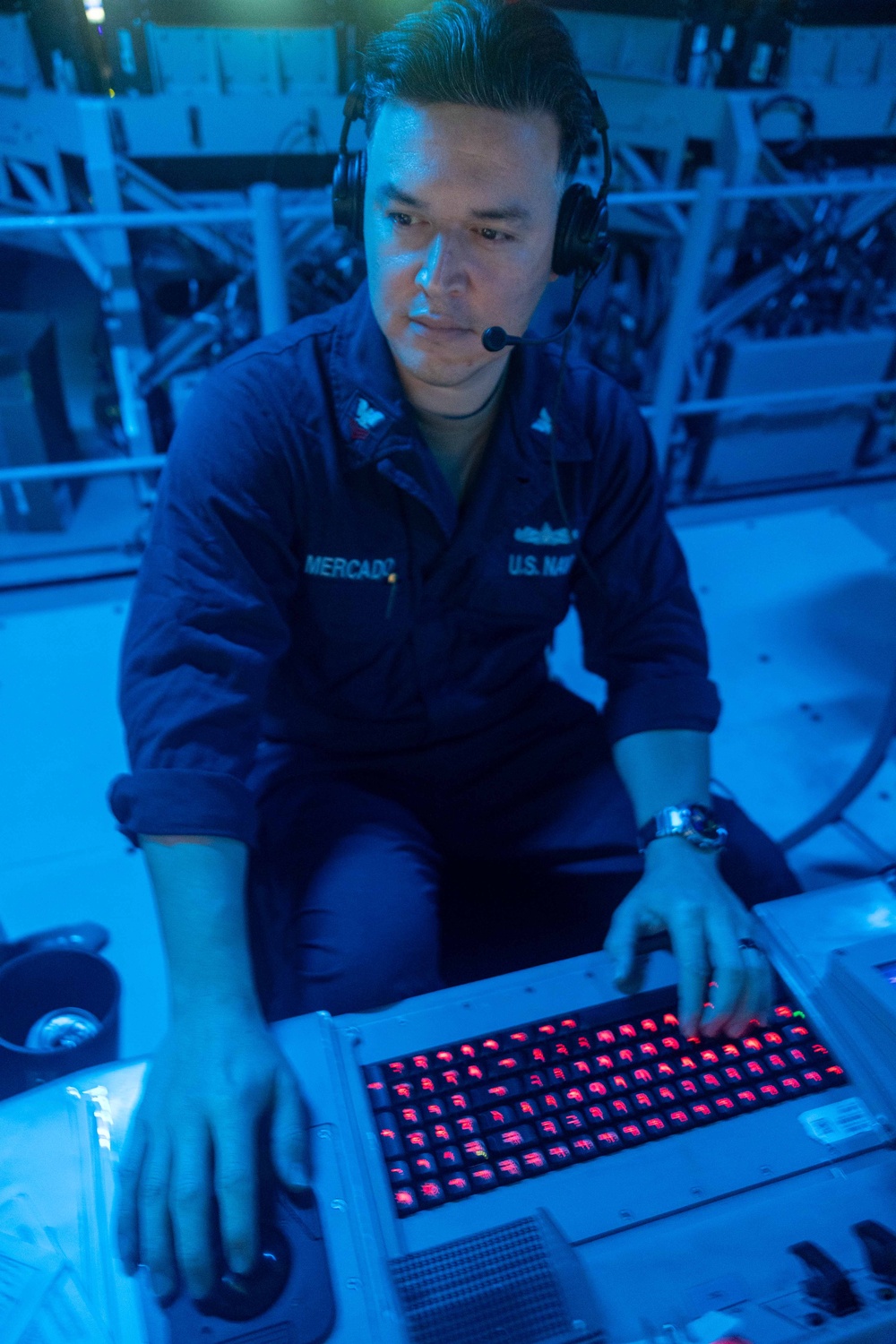 A U.S. Navy sailor identified by the name tape 'MERCADO' is operating what appears to be a specialized military control panel or keyboard with red illuminated keys in a blue-lit environment. The sailor is wearing a headset with a boom microphone and a navy blue coverall uniform, suggesting active duty operations. The setting appears to be aboard a naval vessel, likely in a combat information center or similar operational space.