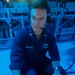 A U.S. Navy sailor identified by the name tape 'MERCADO' is operating what appears to be a specialized military control panel or keyboard with red illuminated keys in a blue-lit environment. The sailor is wearing a headset with a boom microphone and a navy blue coverall uniform, suggesting active duty operations. The setting appears to be aboard a naval vessel, likely in a combat information center or similar operational space.