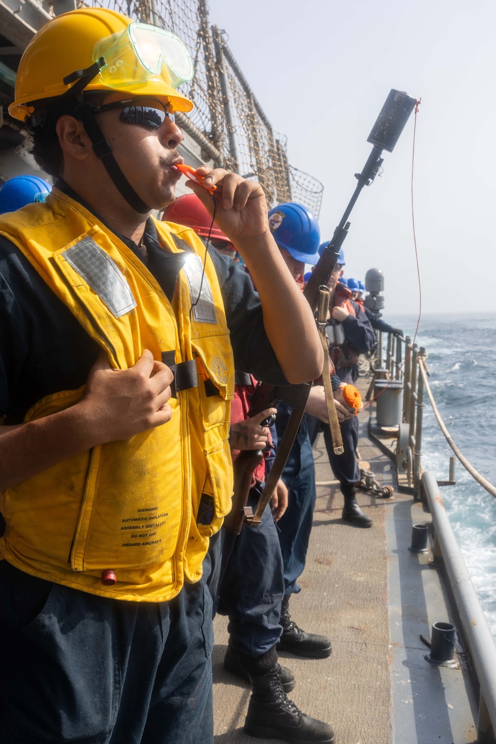 Naval personnel are standing on the deck of a ship alongside a railing with the ocean visible in the background. The foreground sailor wears a yellow hard hat, safety goggles, sunglasses, and a yellow inflatable life vest, and is blowing a whistle while holding what appears to be a line-throwing gun or similar device. Multiple crew members in blue hard hats are lined up behind him in a similar posture, suggesting a replenishment-at-sea (RAS) or man-overboard drill evolution.