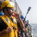 Naval personnel are standing on the deck of a ship alongside a railing with the ocean visible in the background. The foreground sailor wears a yellow hard hat, safety goggles, sunglasses, and a yellow inflatable life vest, and is blowing a whistle while holding what appears to be a line-throwing gun or similar device. Multiple crew members in blue hard hats are lined up behind him in a similar posture, suggesting a replenishment-at-sea (RAS) or man-overboard drill evolution.