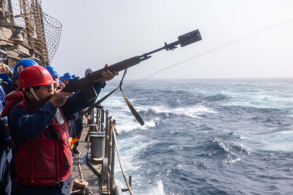 A sailor wearing a red hard hat and life vest is firing a line-throwing rifle from the deck of a naval vessel at sea. A thin line/rope can be seen being projected from the muzzle attachment toward another vessel or target. Other crew members in blue hard hats are visible observing from the ship's rail.