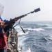 A sailor wearing a red hard hat and life vest is firing a line-throwing rifle from the deck of a naval vessel at sea. A thin line/rope can be seen being projected from the muzzle attachment toward another vessel or target. Other crew members in blue hard hats are visible observing from the ship's rail.