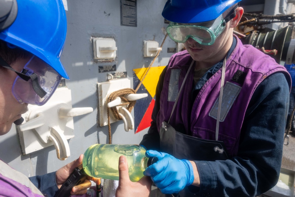 Two workers wearing blue hard hats and safety goggles are examining a glass jar containing a yellow-green liquid, likely a fuel or chemical sample. The setting appears to be an industrial or maritime environment, with equipment panels and cleats visible on a gray metal wall behind them. One worker is wearing blue nitrile gloves and a purple safety vest, suggesting a quality control or sampling procedure.