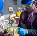 Two workers wearing blue hard hats and safety goggles are examining a glass jar containing a yellow-green liquid, likely a fuel or chemical sample. The setting appears to be an industrial or maritime environment, with equipment panels and cleats visible on a gray metal wall behind them. One worker is wearing blue nitrile gloves and a purple safety vest, suggesting a quality control or sampling procedure.