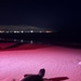 A sea turtle hatchling crawls across a sandy beach at night, illuminated by red/pink artificial lighting. The small hatchling is oriented toward the water, with a distant city skyline visible across the water in the background. Red lighting is commonly used during turtle monitoring to minimize disorientation of hatchlings.