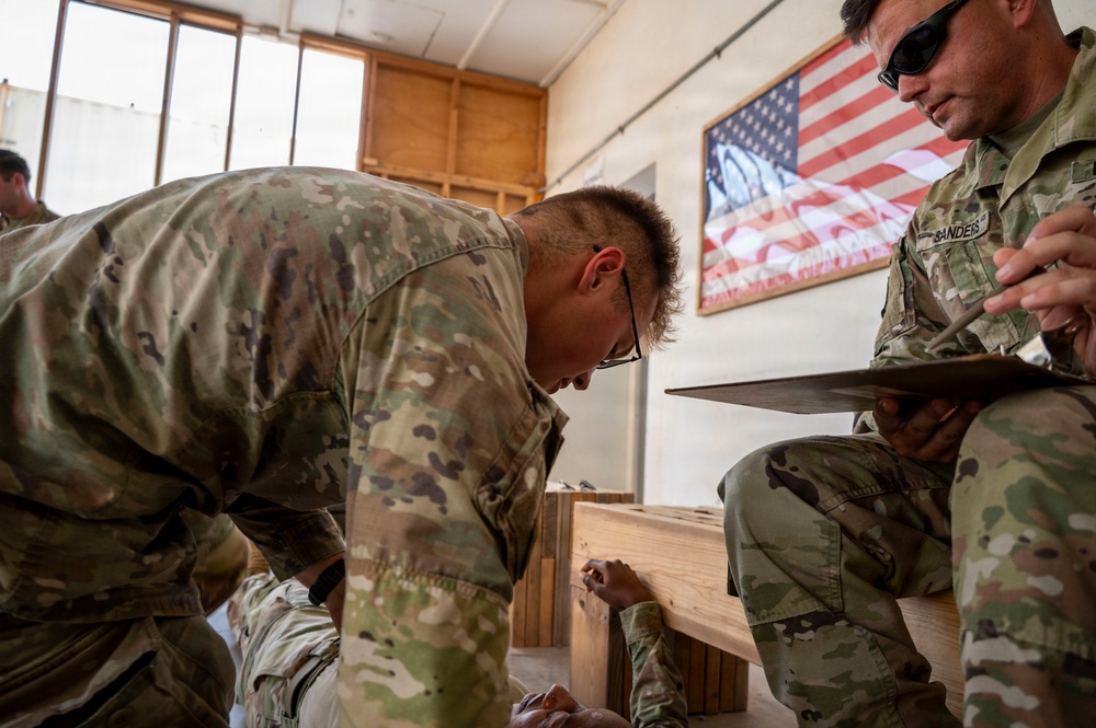 Two U.S. military personnel in MultiCam/OCP camouflage uniforms are engaged in what appears to be a training or assessment activity inside a wooden-structured building. One soldier is leaning over a wooden bench examining something closely, while the other, identified by a name tape reading 'SANDERS,' holds a clipboard and appears to be evaluating or recording observations. An American flag poster is displayed on the wall in the background.
