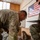 Preparing to Save Lives: CJTF - HOA Medics Conduct MASCAL Exercise Two U.S. military personnel in MultiCam/OCP camouflage uniforms are engaged in what appears to be a training or assessment activity inside a wooden-structured building. One soldier is leaning over a wooden bench examining something closely, while the other, identified by a name tape reading 'SANDERS,' holds a clipboard and appears to be evaluating or recording observations. An American flag poster is displayed on the wall in the background.