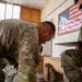 Two U.S. military personnel in MultiCam/OCP camouflage uniforms are engaged in what appears to be a training or assessment activity inside a wooden-structured building. One soldier is leaning over a wooden bench examining something closely, while the other, identified by a name tape reading 'SANDERS,' holds a clipboard and appears to be evaluating or recording observations. An American flag poster is displayed on the wall in the background.