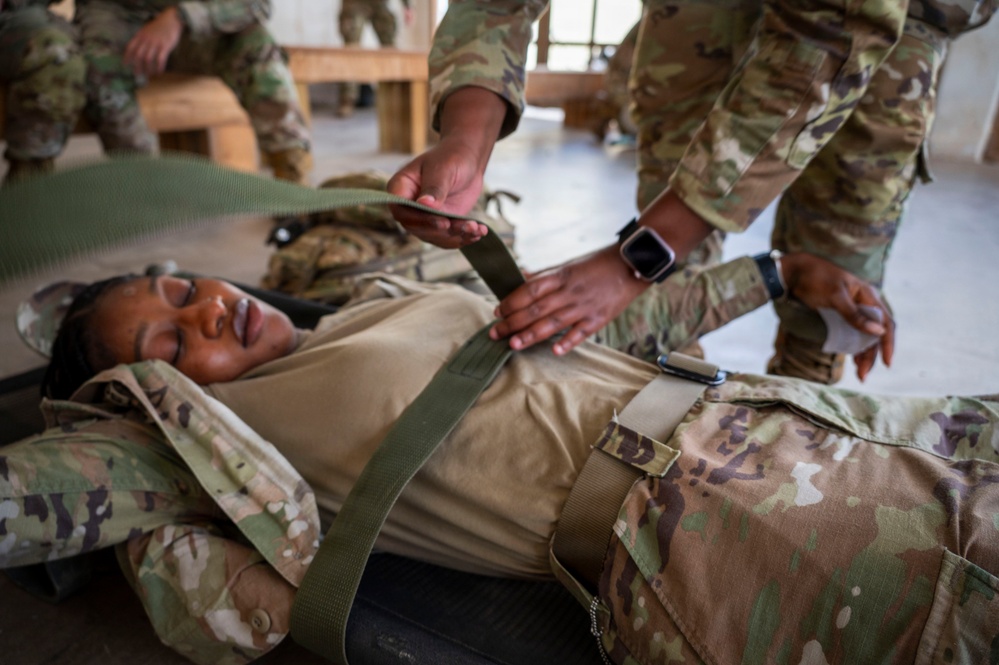 Military personnel in OCP (Operational Camouflage Pattern) uniforms are conducting a medical training exercise. One soldier is lying on what appears to be a litter or stretcher while others practice securing the patient with straps. This appears to be a combat casualty care or tactical combat casualty care (TCCC) training scenario.