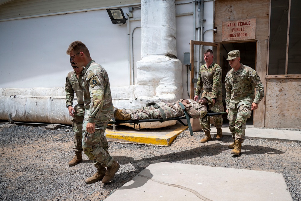 Four military personnel in OCP (Operational Camouflage Pattern) uniforms are carrying a fellow soldier on a litter/stretcher in what appears to be a forward operating base or military installation. The scene takes place outdoors near a building with a 'Male Female Public Restroom' sign, suggesting a base facility. This appears to be a medical evacuation drill or actual casualty transport exercise.