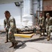 Four military personnel in OCP (Operational Camouflage Pattern) uniforms are carrying a fellow soldier on a litter/stretcher in what appears to be a forward operating base or military installation. The scene takes place outdoors near a building with a 'Male Female Public Restroom' sign, suggesting a base facility. This appears to be a medical evacuation drill or actual casualty transport exercise.