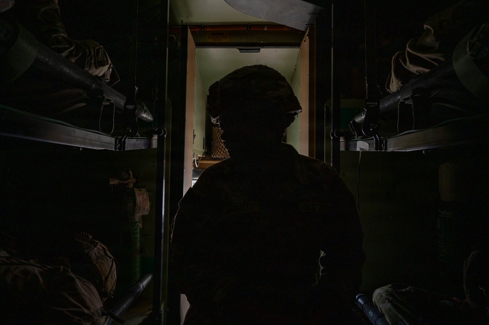 A silhouetted soldier wearing a combat helmet is positioned in a narrow, dark corridor or passageway, likely inside a military vehicle or facility. The figure is backlit by a greenish light source ahead, creating a strong silhouette effect. Military equipment including weapons and gear are visible on racks or shelving on both sides of the corridor.