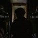 A silhouetted soldier wearing a combat helmet is positioned in a narrow, dark corridor or passageway, likely inside a military vehicle or facility. The figure is backlit by a greenish light source ahead, creating a strong silhouette effect. Military equipment including weapons and gear are visible on racks or shelving on both sides of the corridor.