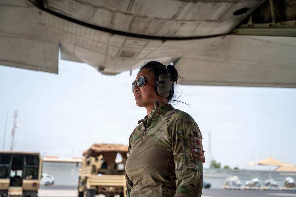 A female military service member in OCP (Operational Camouflage Pattern) uniform stands beneath the wing of a large military aircraft on a flight line. She is wearing hearing protection ear muffs and reflective sunglasses, suggesting active flight line operations. Military vehicles and equipment are visible in the background.