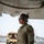 Preparing to Save Lives: CJTF - HOA Medics Conduct MASCAL Exercise A female military service member in OCP (Operational Camouflage Pattern) uniform stands beneath the wing of a large military aircraft on a flight line. She is wearing hearing protection ear muffs and reflective sunglasses, suggesting active flight line operations. Military vehicles and equipment are visible in the background.