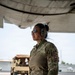 A female military service member in OCP (Operational Camouflage Pattern) uniform stands beneath the wing of a large military aircraft on a flight line. She is wearing hearing protection ear muffs and reflective sunglasses, suggesting active flight line operations. Military vehicles and equipment are visible in the background.