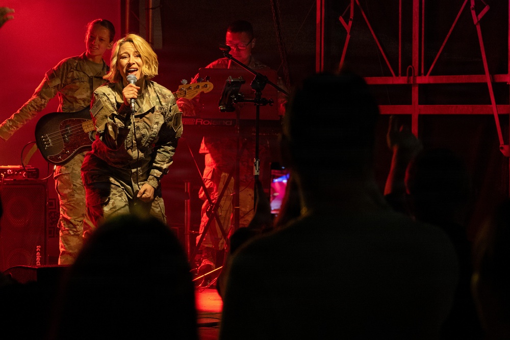 A military band performance is taking place on a stage bathed in red lighting. A female vocalist in camouflage uniform holds a microphone and performs for an audience, while bandmates play bass guitar and keyboard behind her. The scene appears to be an indoor or covered military entertainment event with audience members visible in silhouette in the foreground.