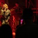 A military band performance is taking place on a stage bathed in red lighting. A female vocalist in camouflage uniform holds a microphone and performs for an audience, while bandmates play bass guitar and keyboard behind her. The scene appears to be an indoor or covered military entertainment event with audience members visible in silhouette in the foreground.