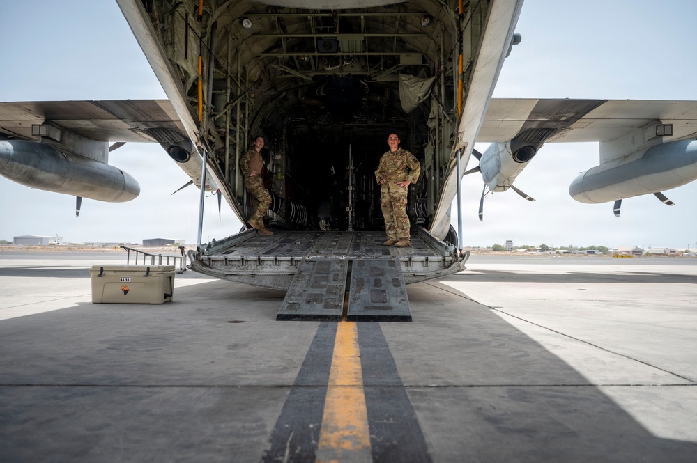 Two military personnel in camouflage uniforms are standing inside the open rear cargo ramp of a large military transport aircraft, likely a C-130 Hercules, on a tarmac. The photo is taken from a ground-level perspective looking directly up the lowered cargo ramp into the aircraft's interior. A white cooler is visible on the tarmac to the left of the ramp.
