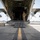Preparing to Save Lives: CJTF - HOA Medics Conduct MASCAL Exercise Two military personnel in camouflage uniforms are standing inside the open rear cargo ramp of a large military transport aircraft, likely a C-130 Hercules, on a tarmac. The photo is taken from a ground-level perspective looking directly up the lowered cargo ramp into the aircraft's interior. A white cooler is visible on the tarmac to the left of the ramp.