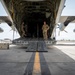 Two military personnel in camouflage uniforms are standing inside the open rear cargo ramp of a large military transport aircraft, likely a C-130 Hercules, on a tarmac. The photo is taken from a ground-level perspective looking directly up the lowered cargo ramp into the aircraft's interior. A white cooler is visible on the tarmac to the left of the ramp.
