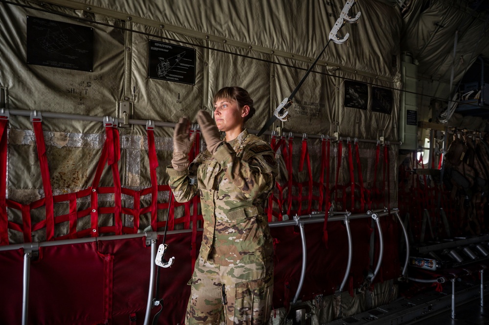 A female military service member in MultiCam/OCP camouflage uniform is standing inside what appears to be a military transport aircraft, likely a C-130 or similar cargo plane. She is reaching upward, possibly inspecting or adjusting equipment along the aircraft's interior wall. The interior features characteristic red troop seating webbing, static line cables, and instructional placards mounted on the fuselage wall.