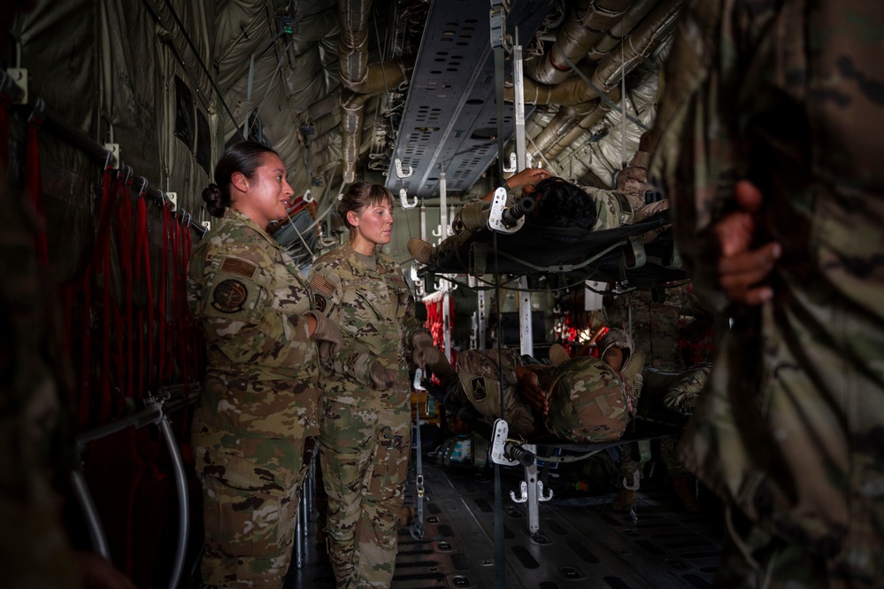 Military personnel in OCP (Operational Camouflage Pattern) uniforms are gathered inside the cargo hold of a large military transport aircraft, likely a C-130 or C-17. The scene appears to depict an aeromedical evacuation training exercise, with litter/stretcher stanchions visible and personnel working with medical transport equipment. Two female service members stand in the foreground observing while others work with the litter stacking system.