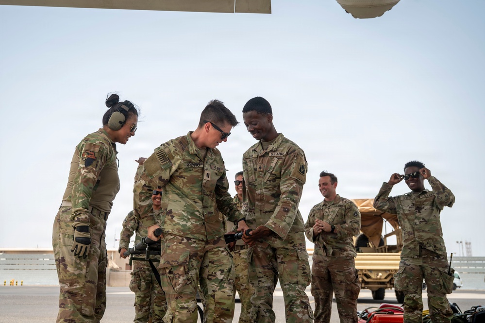 A group of U.S. Army soldiers in OCP (Operational Camouflage Pattern) uniforms are gathered on what appears to be a flight line or tarmac, likely at a forward operating base or airfield in an arid environment. Two soldiers in the foreground appear to be examining or handling a piece of equipment together, while others look on with smiling expressions suggesting a positive or celebratory interaction. The setting and uniforms indicate a deployed military environment.