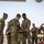 Preparing to Save Lives: CJTF - HOA Medics Conduct MASCAL Exercise A group of U.S. Army soldiers in OCP (Operational Camouflage Pattern) uniforms are gathered on what appears to be a flight line or tarmac, likely at a forward operating base or airfield in an arid environment. Two soldiers in the foreground appear to be examining or handling a piece of equipment together, while others look on with smiling expressions suggesting a positive or celebratory interaction. The setting and uniforms indicate a deployed military environment.