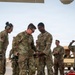 A group of U.S. Army soldiers in OCP (Operational Camouflage Pattern) uniforms are gathered on what appears to be a flight line or tarmac, likely at a forward operating base or airfield in an arid environment. Two soldiers in the foreground appear to be examining or handling a piece of equipment together, while others look on with smiling expressions suggesting a positive or celebratory interaction. The setting and uniforms indicate a deployed military environment.