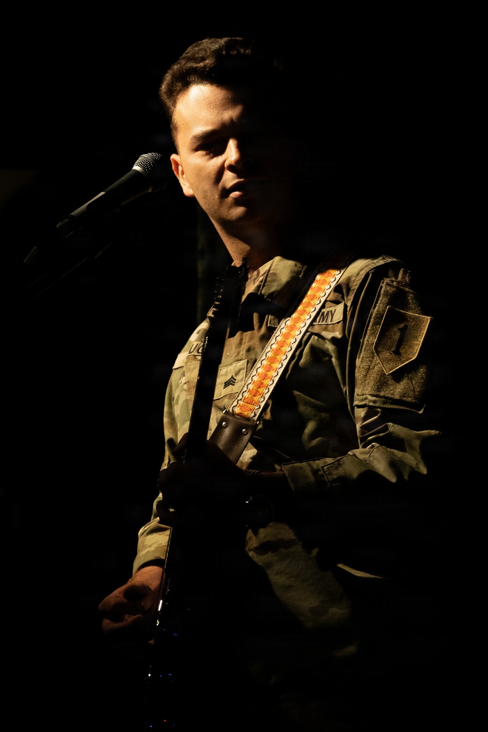A male performer stands on a darkened stage near a microphone, wearing a military-style jacket with visible patches and insignia. He is holding what appears to be a guitar with an orange and tan woven strap crossing his chest. The dramatic low-key lighting illuminates primarily his face and upper body against a near-black background.