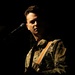 A male performer stands on a darkened stage near a microphone, wearing a military-style jacket with visible patches and insignia. He is holding what appears to be a guitar with an orange and tan woven strap crossing his chest. The dramatic low-key lighting illuminates primarily his face and upper body against a near-black background.
