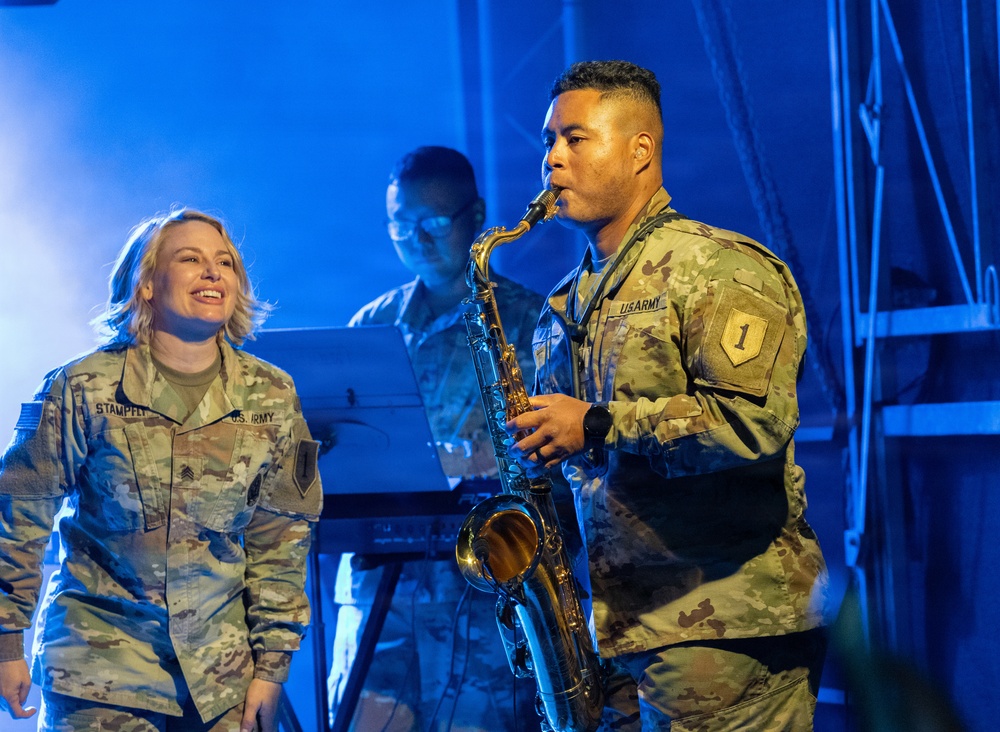 Three U.S. Army soldiers are performing on a stage under blue lighting. A male soldier in the foreground is playing a tenor saxophone while a female soldier to his left watches and smiles. A third soldier is visible in the background, appearing to play a keyboard instrument.