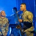 Three U.S. Army soldiers are performing on a stage under blue lighting. A male soldier in the foreground is playing a tenor saxophone while a female soldier to his left watches and smiles. A third soldier is visible in the background, appearing to play a keyboard instrument.