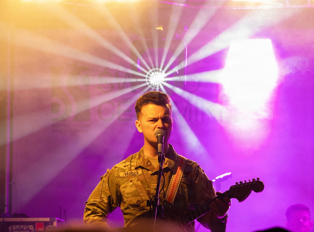 A performer wearing a military camouflage uniform with a name tape reading 'YAUGER' is singing into a microphone while playing an electric guitar on stage. The stage is dramatically lit with purple and golden hues, along with a starburst spotlight effect in the background. The setting appears to be a live concert or military entertainment event.