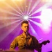 A performer wearing a military camouflage uniform with a name tape reading 'YAUGER' is singing into a microphone while playing an electric guitar on stage. The stage is dramatically lit with purple and golden hues, along with a starburst spotlight effect in the background. The setting appears to be a live concert or military entertainment event.
