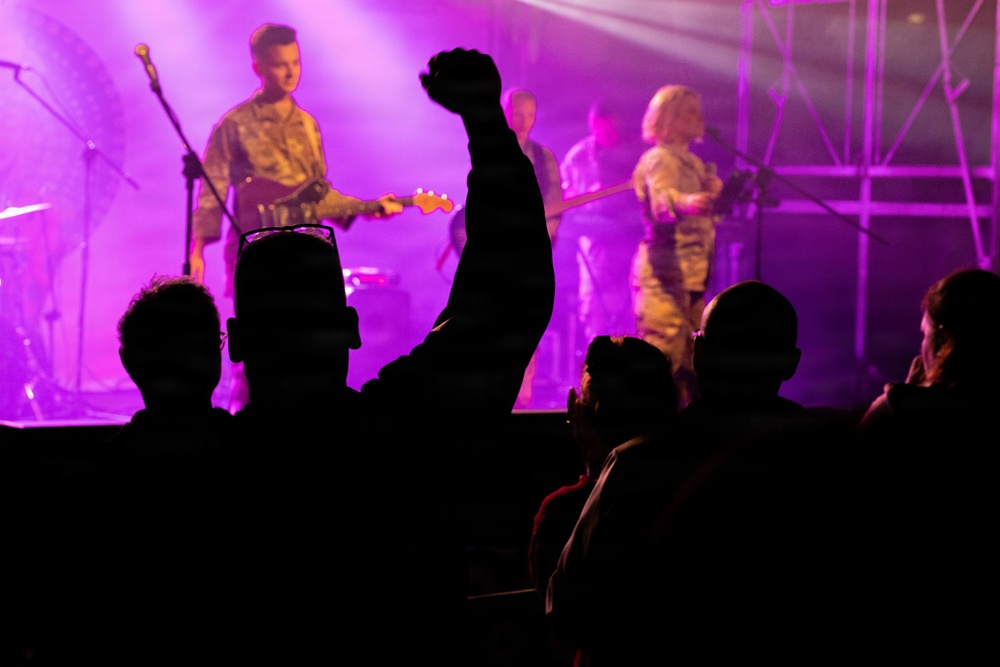 A live music concert is taking place on a purple/pink-lit stage with multiple performers visible. The audience is silhouetted in the foreground, with one person raising a fist in the air in an enthusiastic gesture. The performers appear to be wearing military-style camouflage uniforms while playing instruments.
