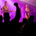 A live music concert is taking place on a purple/pink-lit stage with multiple performers visible. The audience is silhouetted in the foreground, with one person raising a fist in the air in an enthusiastic gesture. The performers appear to be wearing military-style camouflage uniforms while playing instruments.