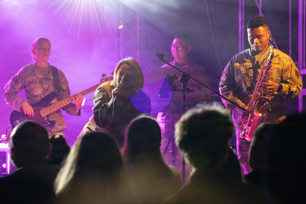 Military personnel in camouflage uniforms are performing a live musical concert on a lit stage with purple lighting effects. The band includes a vocalist, bass guitarist, saxophonist, and at least one additional musician in the background. An audience is visible in silhouette in the foreground watching the performance.