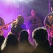 Military personnel in camouflage uniforms are performing a live musical concert on a lit stage with purple lighting effects. The band includes a vocalist, bass guitarist, saxophonist, and at least one additional musician in the background. An audience is visible in silhouette in the foreground watching the performance.