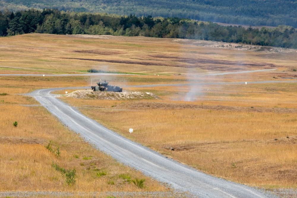 A military armored vehicle is positioned on an elevated mound beside a gravel road in an open training range or firing range. Smoke or dust is visible rising near the vehicle, suggesting recent weapons discharge or movement activity. The surrounding landscape consists of dry golden grassland with a treeline visible in the background.