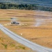 A military armored vehicle is positioned on an elevated mound beside a gravel road in an open training range or firing range. Smoke or dust is visible rising near the vehicle, suggesting recent weapons discharge or movement activity. The surrounding landscape consists of dry golden grassland with a treeline visible in the background.