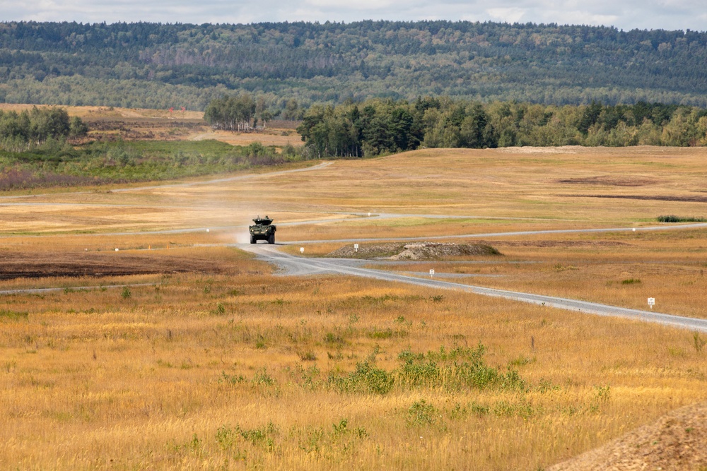A military wheeled armored vehicle is traveling along a gravel road through an open, arid training range landscape. The vehicle is raising a visible dust cloud behind it as it moves forward. The terrain is characterized by dry golden grassland flanked by tree lines and rolling hills in the background.