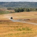 A military wheeled armored vehicle is traveling along a gravel road through an open, arid training range landscape. The vehicle is raising a visible dust cloud behind it as it moves forward. The terrain is characterized by dry golden grassland flanked by tree lines and rolling hills in the background.