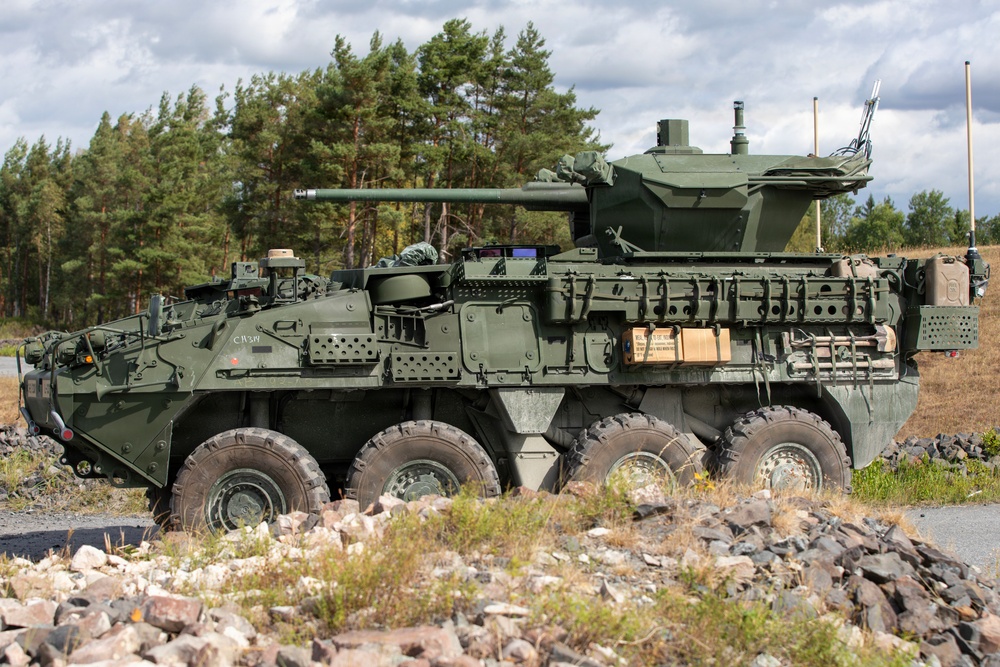 The image shows a military wheeled armored fighting vehicle, consistent with a Stryker-family or LAV-type 8x8 infantry carrier variant fitted with a medium-caliber cannon turret. The vehicle is painted in olive drab/military green and is positioned on rocky terrain near a treeline. The setting appears to be a training area or exercise environment in a northern European or similar temperate forested region.