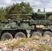 The image shows a military wheeled armored fighting vehicle, consistent with a Stryker-family or LAV-type 8x8 infantry carrier variant fitted with a medium-caliber cannon turret. The vehicle is painted in olive drab/military green and is positioned on rocky terrain near a treeline. The setting appears to be a training area or exercise environment in a northern European or similar temperate forested region.